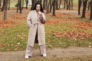 vrouw in een grijs jas wandelen in de herfst park met een kop van heet drinken foto