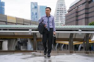 Aziatische zakenman lopen en bedrijf aktetas met zakelijke kantoorgebouwen op de achtergrond van de stad foto