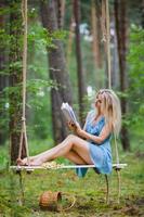 mooi blond vrouw in kort blauw jurk lezing een boek Aan een touw schommel foto