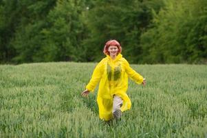 een jong meisje in een geel regenjas loopt door een groen tarwe veld- foto