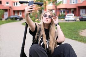 een tiener- meisje in zonnebril duurt een selfie Aan een scooter foto