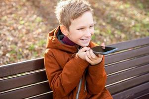een jongen records een stem bericht en lacht terwijl zittend Aan een park bank in herfst foto
