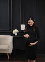 een zwanger vrouw vervelend een zwart jurk in voorkant van sofa en lamp met donker achtergrond, iets groot is komt eraan foto
