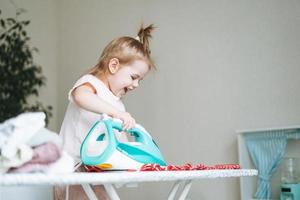 schattig weinig baby meisje spelen en strijken kleren met een ijzer Bij huis foto