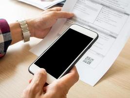 dichtbij omhoog hand- Holding slim telefoon naar scannen qr code van factuur Aan papieren voor de rekeningen. online Bill betaling concept foto