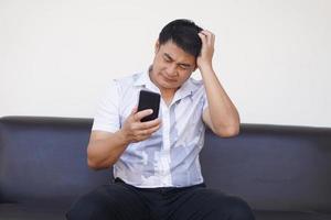 Aziatisch Mens zit Aan bank, houden smartphone, voelt hoofdpijn. van streek, verdrietig. concept, weten slecht nieuws van sociaal media online. gevoel en emotie uitdrukking. depressie, mislukking, teleurstelling, gebroken hart foto