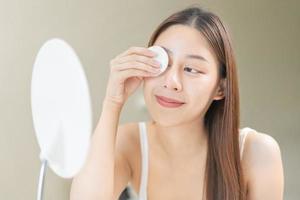 gelukkig schoonheid, mooi Aziatisch jong vrouw, meisje op zoek in naar spiegel, Holding katoen kussen, toepassen gelaats veeg Aan haar gezicht, Verwijderen bedenken voordat douche in badkamer, huid zorg Aan wit achtergrond. foto