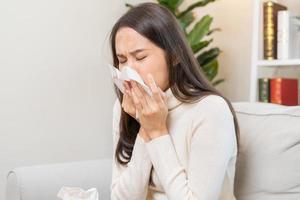 ziek, coronavirus covid-19 Aziatisch jong vrouw, meisje hoofdpijn onder deken hebben een koorts, griep en gebruik weefsels papier niezen neus, vloeibaar zittend Aan sofa bed Bij huis. Gezondheid zorg Aan virus persoon. foto