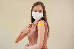 portret van Gezondheid zorg Aziatisch jong vrouw vervelend gezicht masker beschermen, krijgen vaccin van covid19, influenza in gewoontjes, tonen arm Aan geel verband, gips, geïsoleerd Aan natuur achtergrond, kopiëren ruimte foto