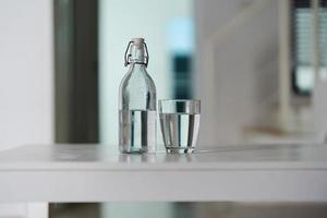 een glas van schoon drinken water en een water fles Aan een wit houten tafel foto