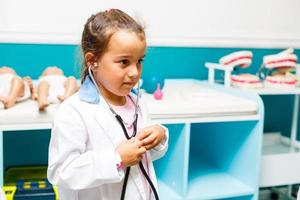 een weinig schattig glimlachen meisje vervelend een dokter uniform met stethoscoop in een ziekenhuis foto