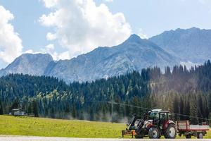 oogsten Bijsnijden door rood trekker in oostenrijks alpine groen berg velden met pluizig wolken Aan lucht foto