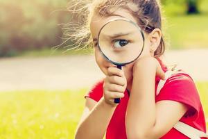 positief vrolijk weinig meisje op zoek door een vergroten glas buitenshuis foto