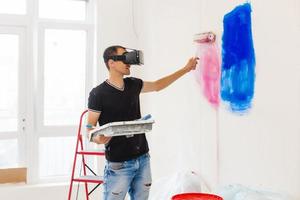 jong Mens met donker haar- is schilderij de muur in wit in zijn appartement. in zijn handen hij houdt een verf rol en verf borstel. kan niet besluiten wat naar Kiezen. foto