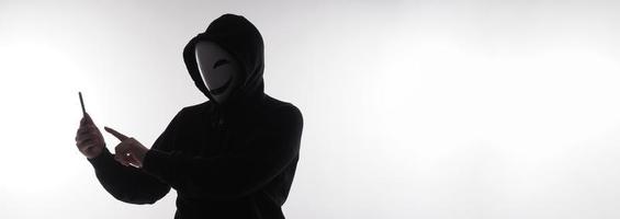 hacker anoniem en gezicht masker met smartphone in hand. Mens in zwart kap overhemd Holding en gebruik makend van mobiel telefoon Aan wit achtergrond. staan voor cyber misdrijf gegevens hacken of stelen persoonlijk gegevens concept foto