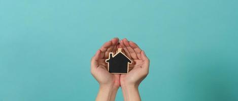 klein hout huis in handen staan voor concepten zo net zo huis zorg familie liefde echt landgoed behuizing onderdak verzekering en hypotheek. handen Holding klein model- huis geïsoleerd Aan blauw groen studio achtergrond. foto