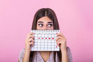 portret van een grappig jong meisje in schuilplaats achter een menstruatie- periodes kalender en op zoek weg Bij kopiëren ruimte geïsoleerd over- roze achtergrond. vrouw periode kalender foto