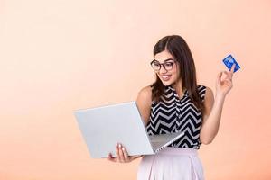 gelukkig mooi brunette vrouw in trui met bril en laptop computer Holding credit kaart en op zoek weg over- roze achtergrond foto