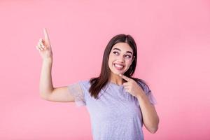 portret van brunette vrouw met lang haar- in eenvoudig t-shirt verheugt en richten vinger Bij copyspace geïsoleerd over- roze achtergrond foto