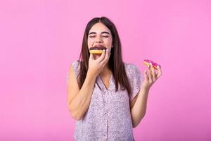 dichtbij omhoog portret van een tevreden mooi meisje aan het eten donuts geïsoleerd over- roze achtergrond. jong gelukkig Kaukasisch meisje aan het eten chocola donuts in voorkant van roze muur foto