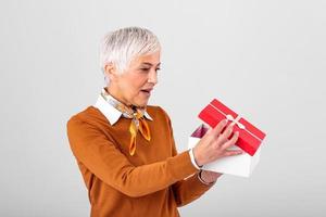 portret van een gelukkig glimlachen volwassen vrouw opening een Kerstmis geschenk doos geïsoleerd over- grijs achtergrond. opgewonden senior gewoontjes vrouw Holding Cadeau doos foto