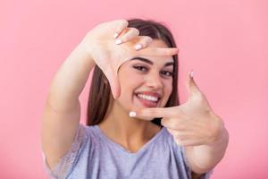 gelukkig jong vrouw maken kader gebaar met vingers over- roze achtergrond. portret van een gelukkig glimlachen vrouw framing haar gezicht met haar handen geïsoleerd foto