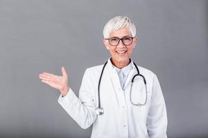 dokter senior vrouw, medisch professioneel richten in Rechtsaf richting geïsoleerd over- grijs achtergrond. vrouw volwassen dokter met stethoscoop foto