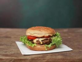 groot hamburger leugens Aan ambacht wit papier tegen houten tafel. een sappig groen salade blad en een rood tomaat liggen in de buurt de hamburger foto