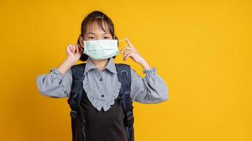 jong Aziatisch meisje dat een gezichtsmasker en een rugzak draagt die de camera met gele achtergrond bekijkt