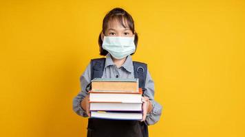 jong thais meisje dat gezichtsmasker draagt en een stapel boeken in een studio met gele achtergrond houdt foto