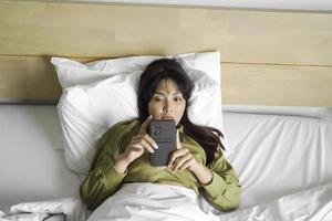 een jong Aziatisch vrouw is gevoel echt en gefocust Aan Holding haar smartphone in hand- terwijl aan het liegen Aan de bed. foto