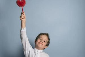 een schattig Kaukasisch jongen in een wit overhemd houdt een pluizig rood hart in zijn verheven hand- foto