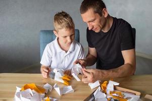 een schattig jongen is zittend met zijn vader Bij de tafel en verzamelen origami. lijm de onderdelen foto
