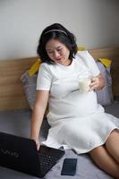 gelukkig jong Aziatisch zwanger vrouw drinken melk Aan bed Bij huis terwijl Holding haar buik en denken foto
