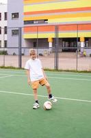een tiener- jongen staat Aan een groen veld- in de school- werf met een voetbal bal in opleiding foto