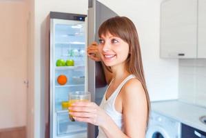jong brunette meisje met een glas van sap in de buurt de koelkast foto