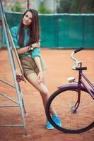 mooi jong meisje met longboard en fiets staand Aan de tennis rechtbank foto