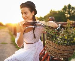 jong mooi vrouw zittend Aan haar fiets met bloemen Bij zonsondergang foto