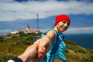 volgen me - gelukkig jong vrouw in een rood hoed en met een rugzak achter haar terug trekken jongens hand. hand- in hand- wandelen naar de vuurtoren foto