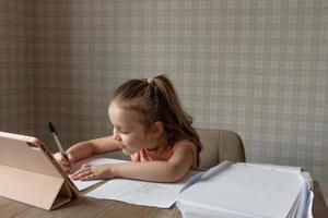 een weinig meisje zit Bij een tafel in voorkant van haar een tablet en een map van wit papier, de kind trekt en kopieën van de tablet, online tekening lessen foto