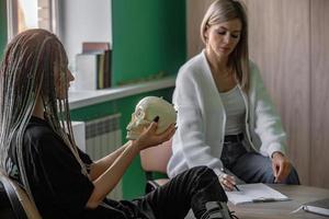 een vrouw psycholoog werken in haar kantoor met een jong cliënt met dreadlocks Aan haar hoofd, wie is Holding een mock-up van een schedel. problemen van generatie z in leven, in communicatie. foto