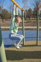 een sport- meisje van negen jaren is verloofd in een horizontaal bar in de park terwijl wandelen Aan een zonnig dag foto