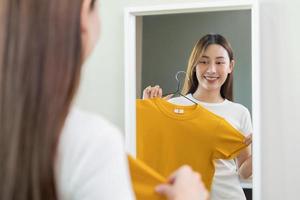 keuze van kleren, niets naar dragen. aantrekkelijk Aziatisch jong vrouw, meisje op zoek in spiegel, proberen Aan verschijnen, kiezen jurk, kleding Aan hanger in garderobe Bij huis. beslissen blouse wat naar zetten Aan welke een foto