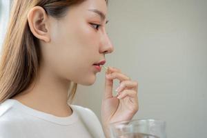 dieet supplement, Aziatisch jong vrouw, meisje hand- Holding geel medisch tablet of pil, nemen vis olie of vitamine c, d voor behandeling voor huid, haar- met glas van zuiver water Bij huis, schoonheid gezondheidszorg. foto