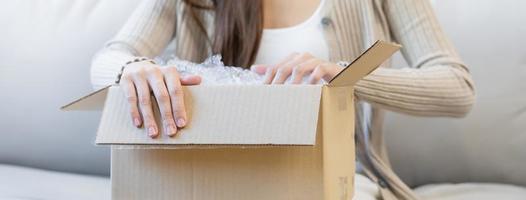 gelukkig Aziatisch jong vrouw, meisje klant zittend Aan sofa Bij huis, hand- in opening en uitpakken karton doos karton pakket na buying bestellen Cadeau, boodschappen doen online, levering onderhoud concept. foto