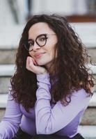 mooi jong vrouw met brunette gekruld haar, portret in oog bril genieten van de zon in de stad. foto