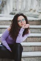 mooi jong vrouw met brunette gekruld haar, portret in oog bril genieten van de zon in de stad. foto