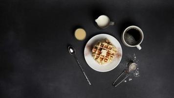 top visie van smakelijk wafels bord, karamel saus, koffie beker, melk, dessert lepel, zeef Aan een zwart beton achtergrond foto