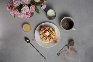 top visie van smakelijk wafels bord, karamel saus, koffie beker, melk, dessert lepel, zeef, roze bloemen Aan een grijs beton achtergrond foto