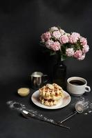 smakelijk wafels bord, karamel saus, koffie beker, melk, dessert lepel, zeef, roze bloemen Aan een zwart beton achtergrond foto